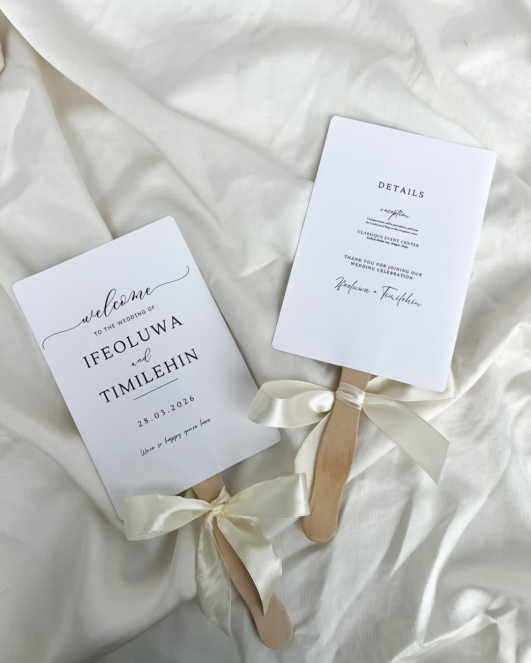 Two minimalist white wedding program fans with wooden handles and cream satin bows.