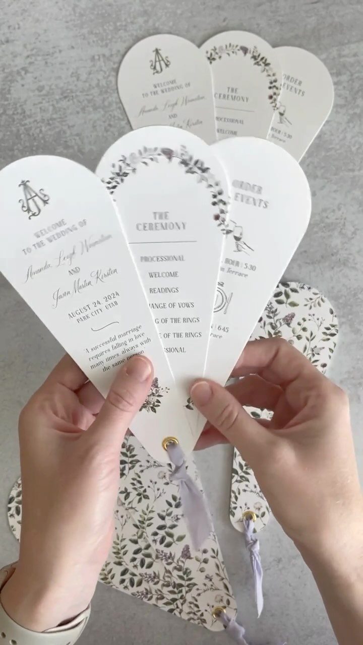 White folding wedding program fans with floral accents held by two hands.