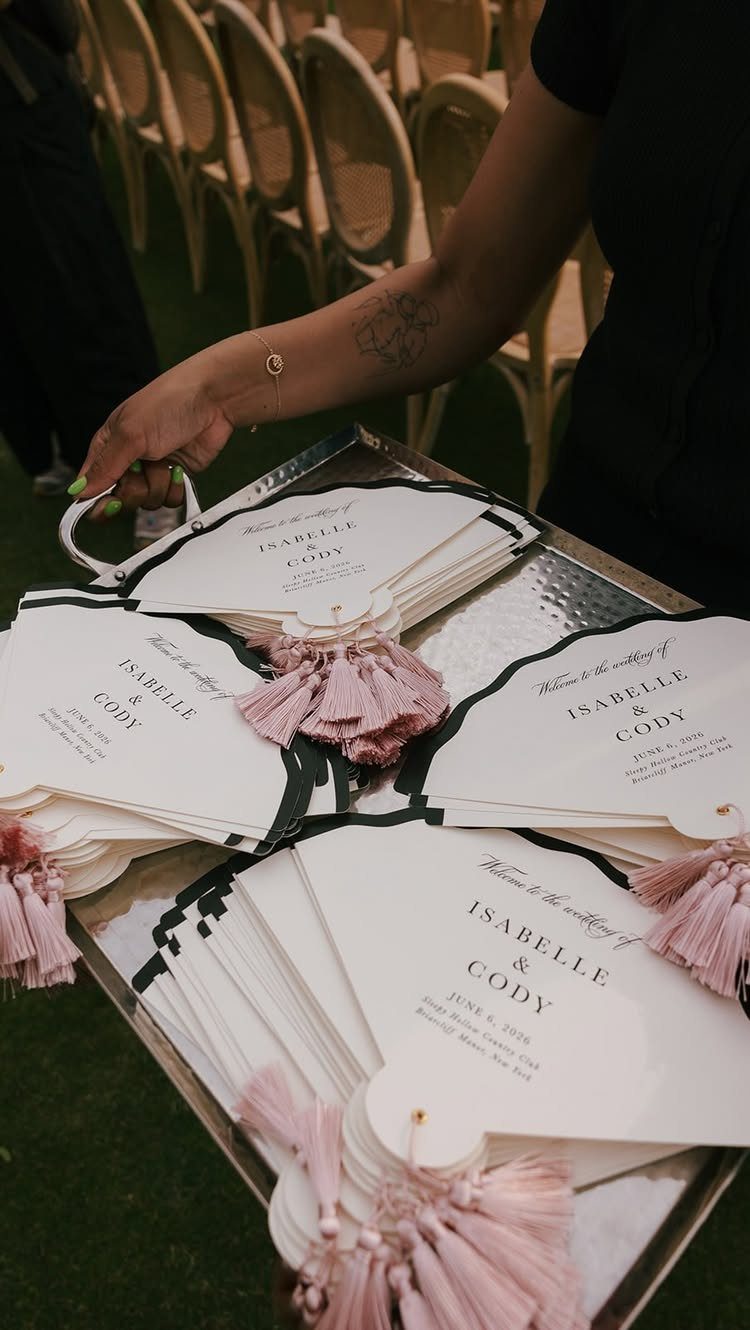 White paddle-style wedding program fans with pink tassels on a silver tray.