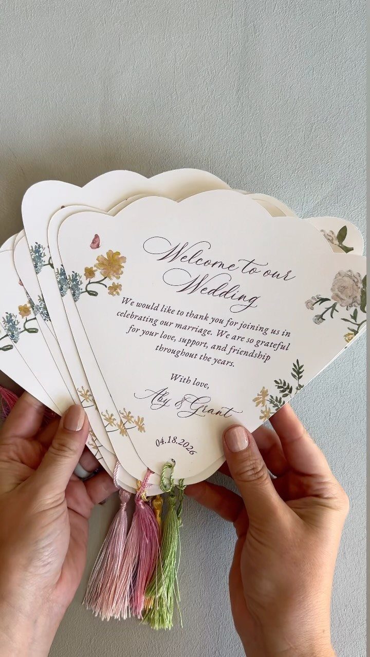 White cardstock wedding program fans with floral designs and colorful hanging tassels.