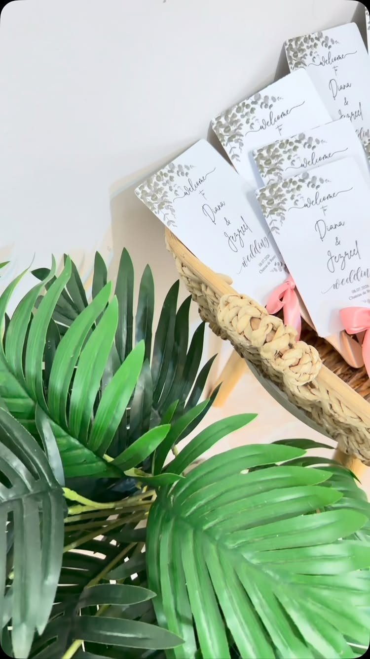 Wedding program fans with floral designs in a woven basket with palm leaves.