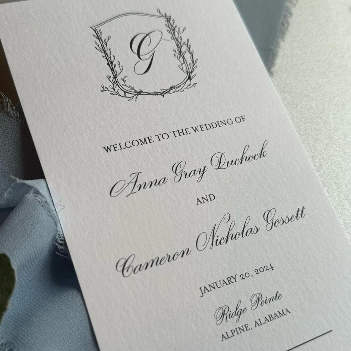 Wedding program card with elegant black calligraphy and a floral crest on white paper.