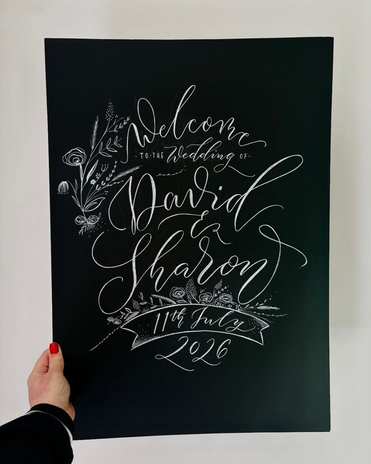 Black chalkboard wedding welcome sign with white calligraphy and floral accents.