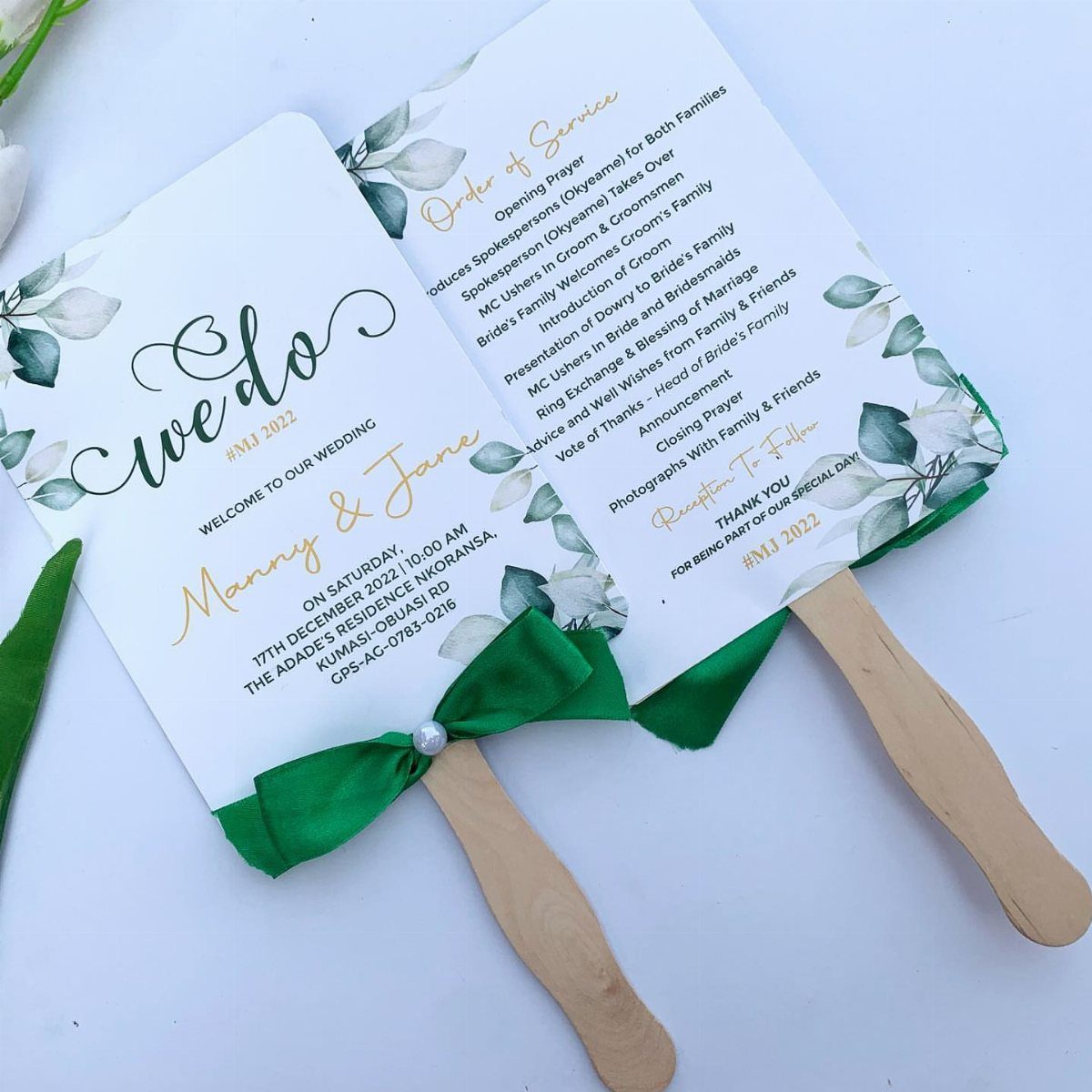 Two white wedding fan programs with wooden handles, green ribbons, and floral designs.