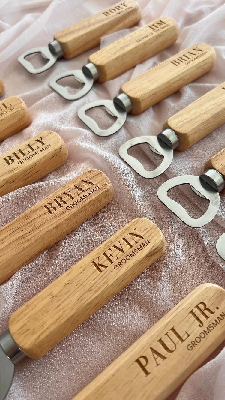 Personalized wooden bottle openers with engraved groomsmen names on pink draped fabric.