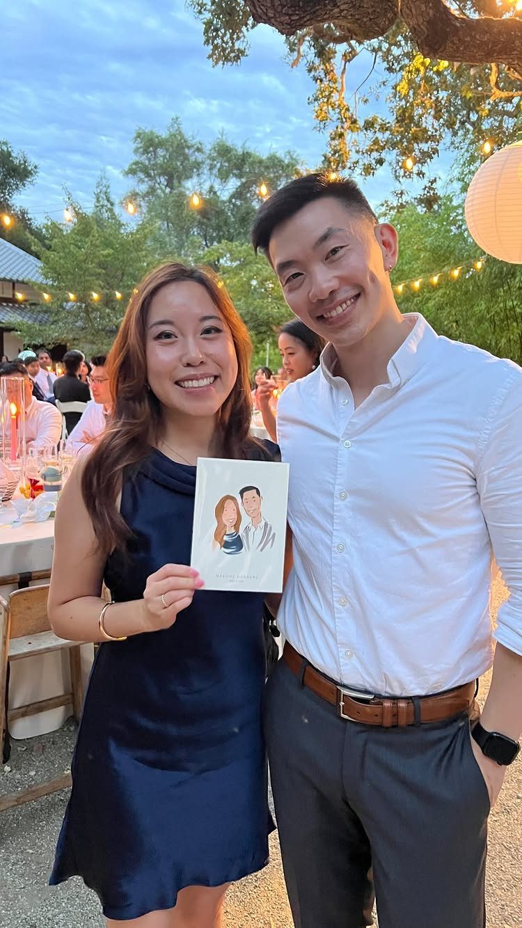 Smiling couple holding a small custom illustration of themselves at an outdoor wedding.