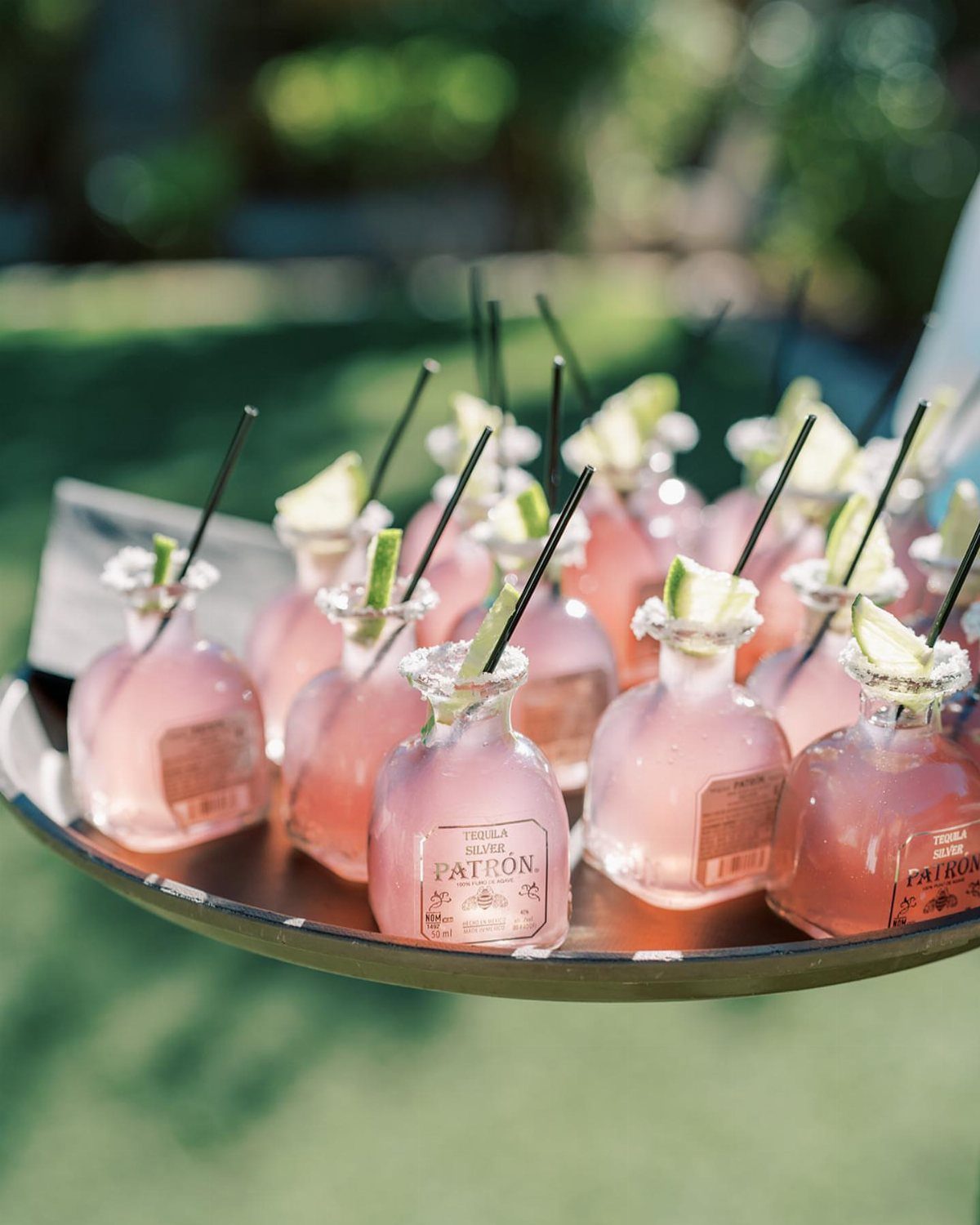 Small round glass bottles with pink cocktails, black straws, and apple slices on a tray.