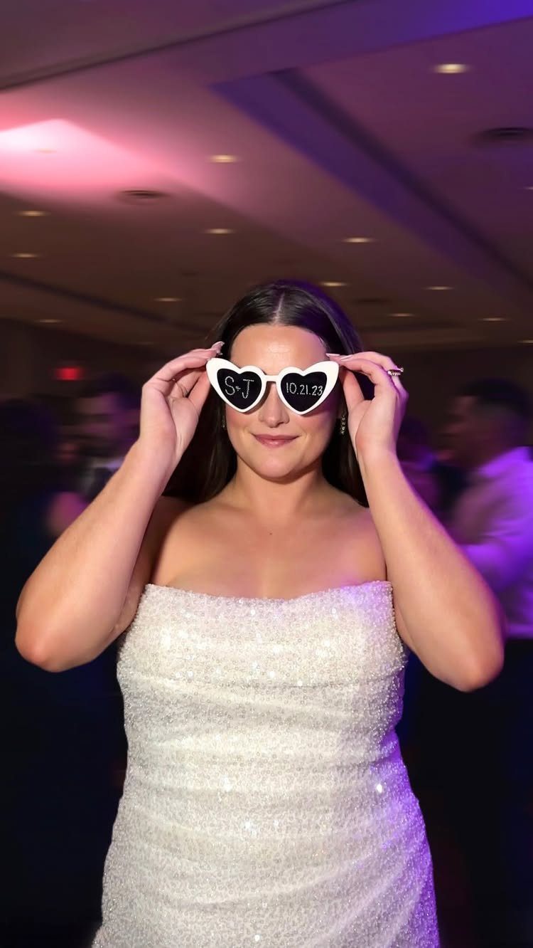 Bride in a white sequined dress wearing heart-shaped sunglasses at a wedding reception.
