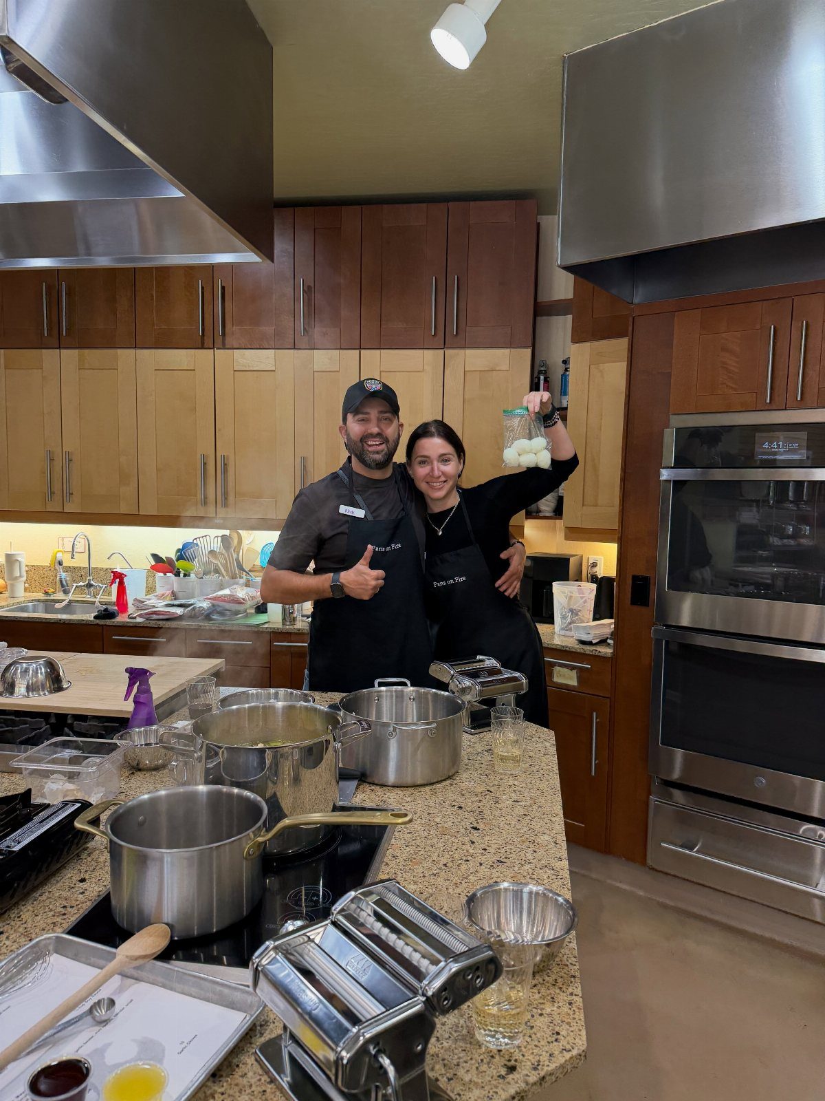 Couple in black aprons posing in a kitchen with pasta making equipment.