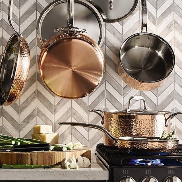 Copper cookware hanging and on a stove in a modern kitchen with herringbone tiles.