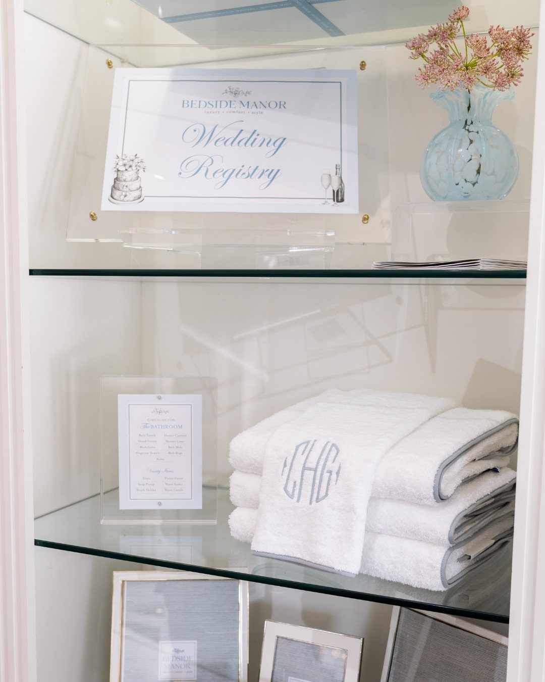 White monogrammed towels stacked on a glass shelf next to a wedding registry sign.