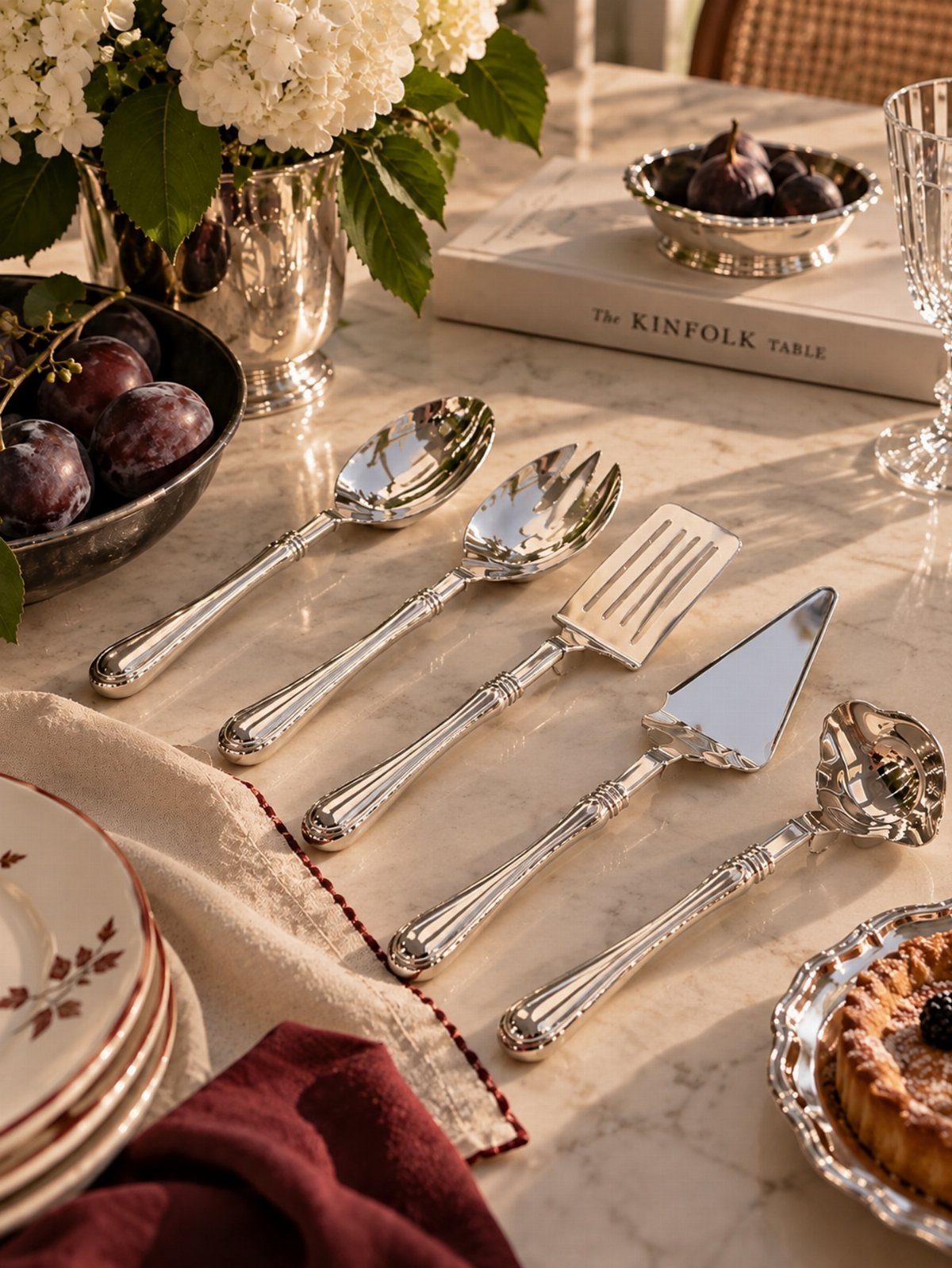 Silver serving utensils arranged on a white marble table with fruit and flowers.