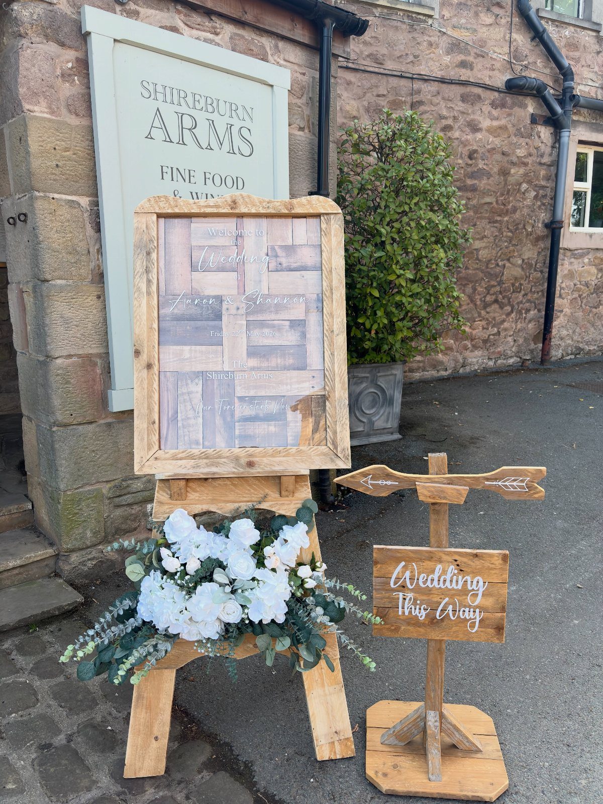 Wooden directional wedding sign and framed welcome sign with white floral arrangements outdoors.