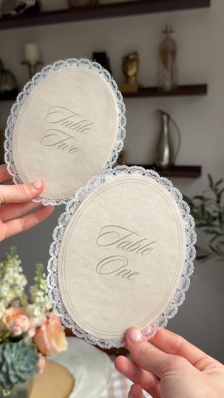 Oval cream table number signs with white lace borders held by two hands.