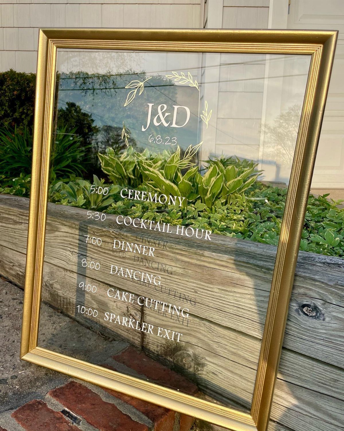Gold framed wedding timeline sign listing event schedule outdoors near green plants.