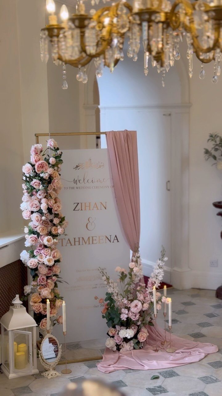 Wedding welcome sign with gold text, pink flowers, and draped fabric under a chandelier.