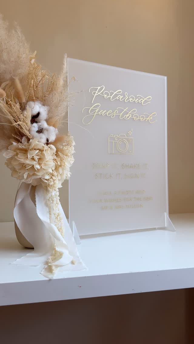 White acrylic guestbook sign with gold text next to dried floral arrangement.
