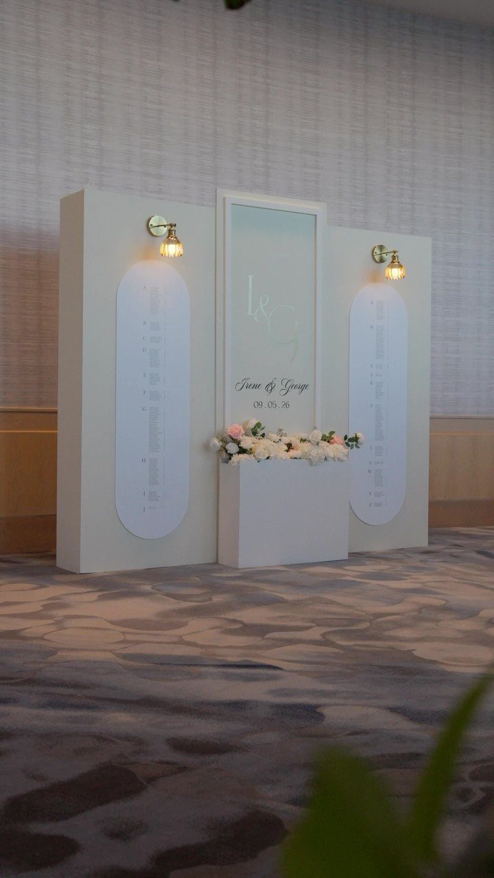 White three-panel wedding seating chart with gold lights and floral arrangement on pedestal