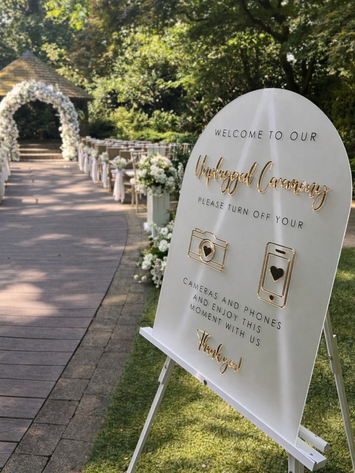 White arched unplugged ceremony sign with gold text on a white easel outdoors.