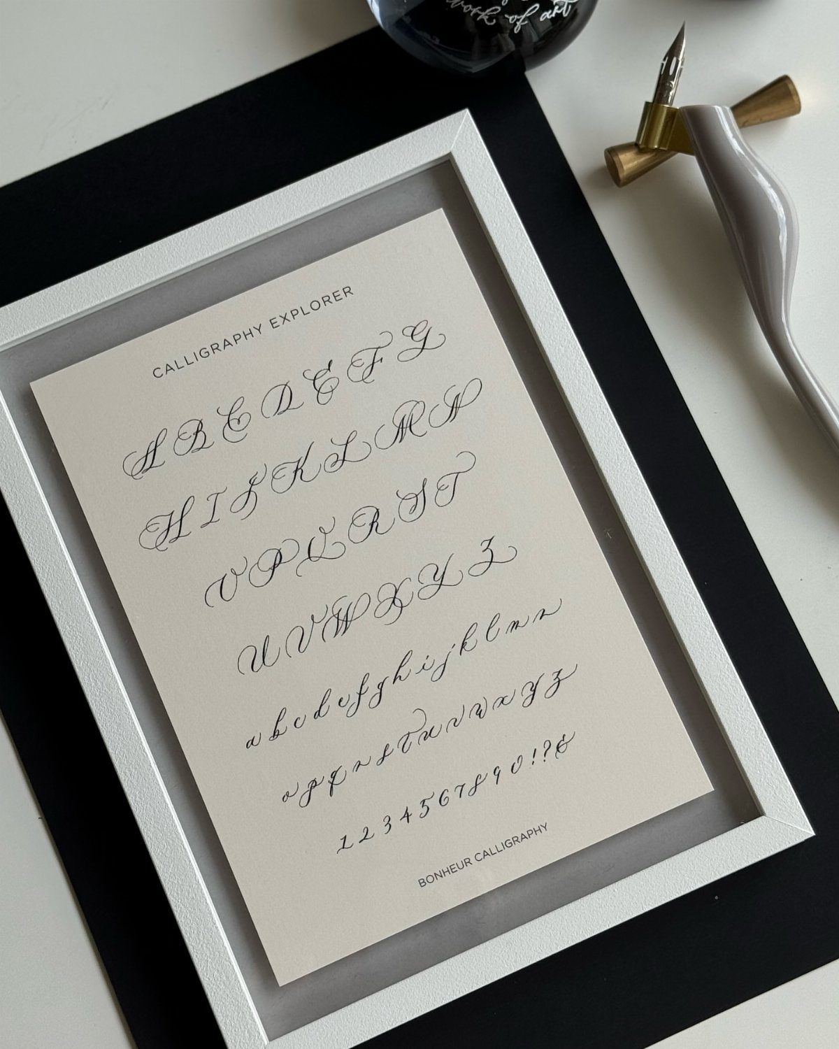 Calligraphy alphabet and numbers in elegant script displayed in a white frame.