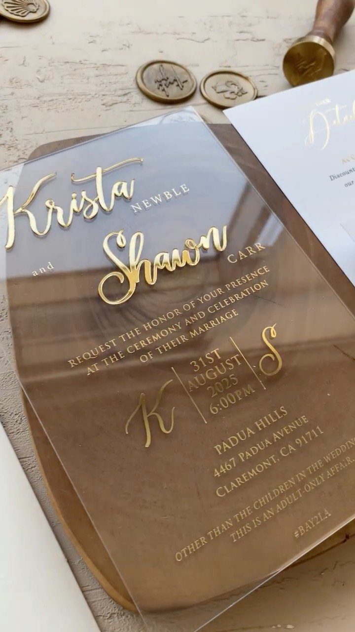 Acrylic wedding invitation with gold lettering resting on a brown wooden board.
