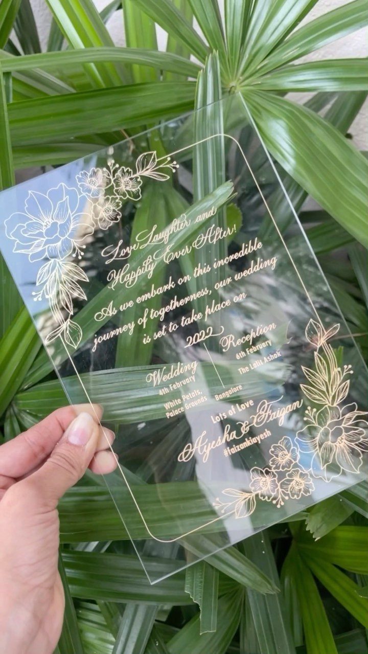 Acrylic wedding invitation with gold floral details held over green palm leaves