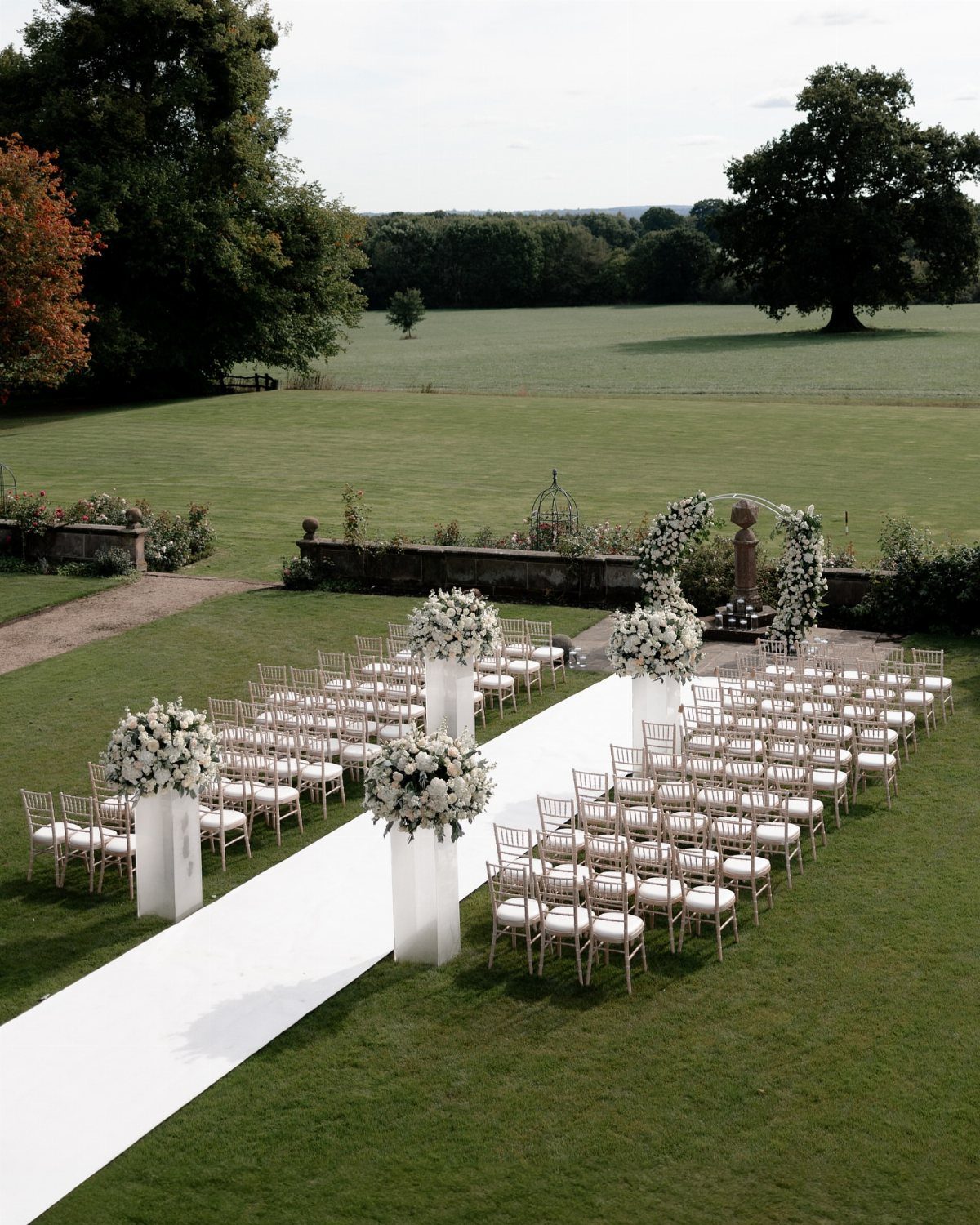 Outdoor wedding ceremony setup with white chairs, aisle runner, and floral arch.