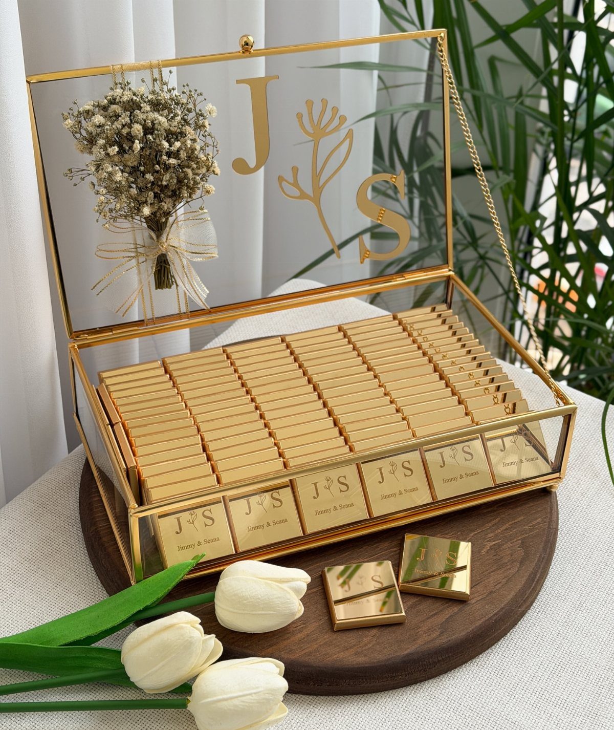 Gold-wrapped chocolates in a glass display box with initials and dried flowers.