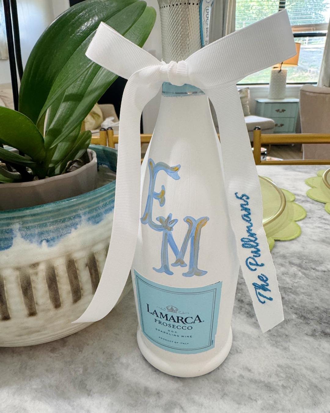 White painted prosecco bottle with blue initials and a personalized white ribbon bow.